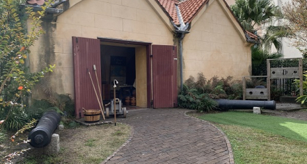 Image Of A Brick Path That Leads To A Light Tan Building With A Terracotta Tile Roof And A Wide Open Doorway With Brown Doors.